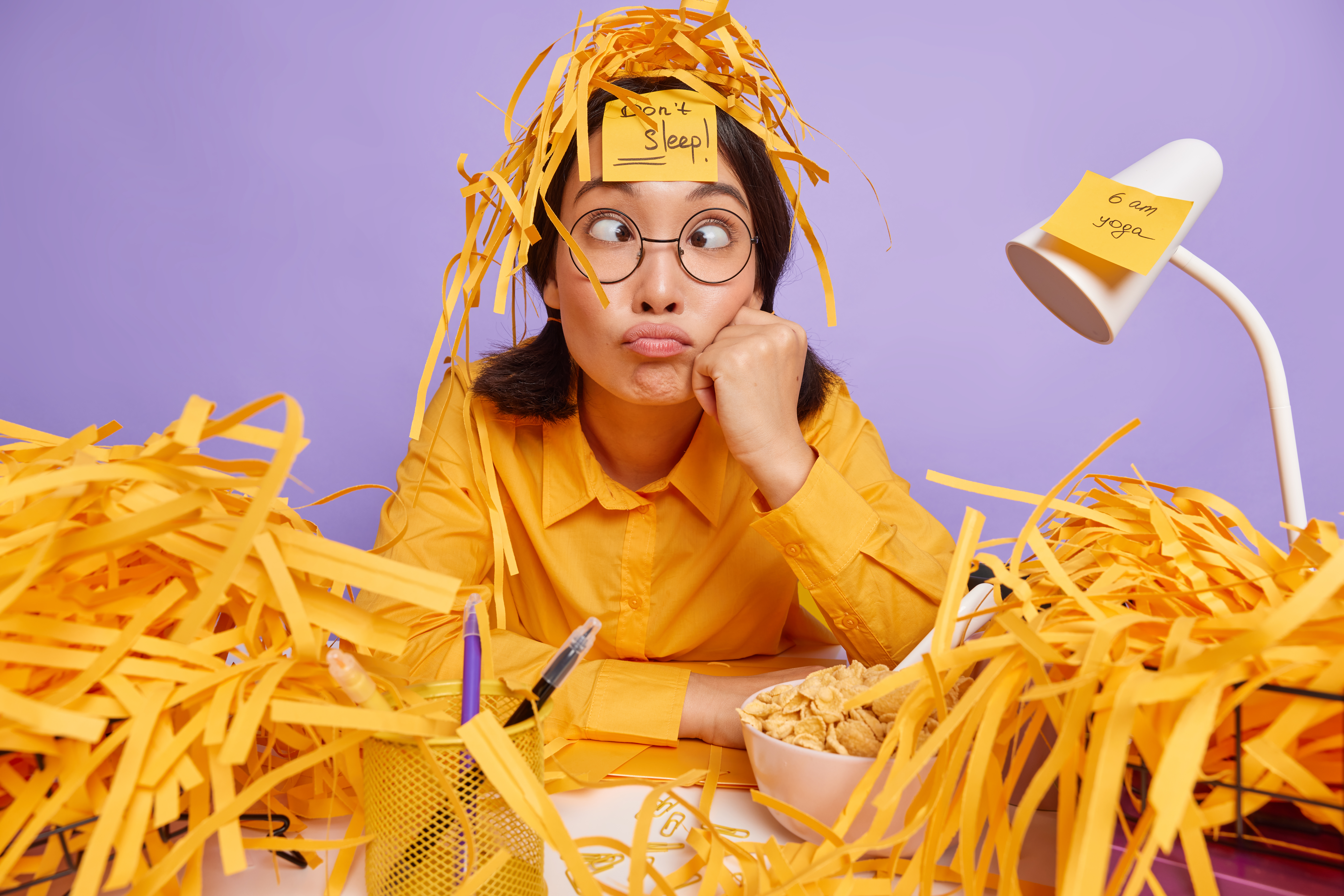 Funny schoolgirl prepares for test at home has sleepless night works late hours makes grimace has busy working schedule surrounded by paper wastes isolated over purple background. Deadline and work